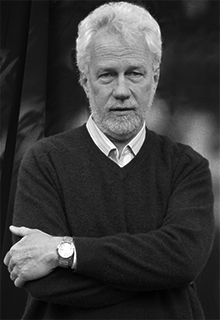 A black and white image of a white man wearing a sweater and a watch crossing his arms