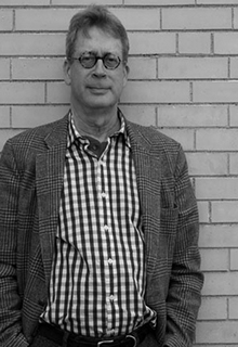 A black and white image of a white man standing in front of a brick wall
